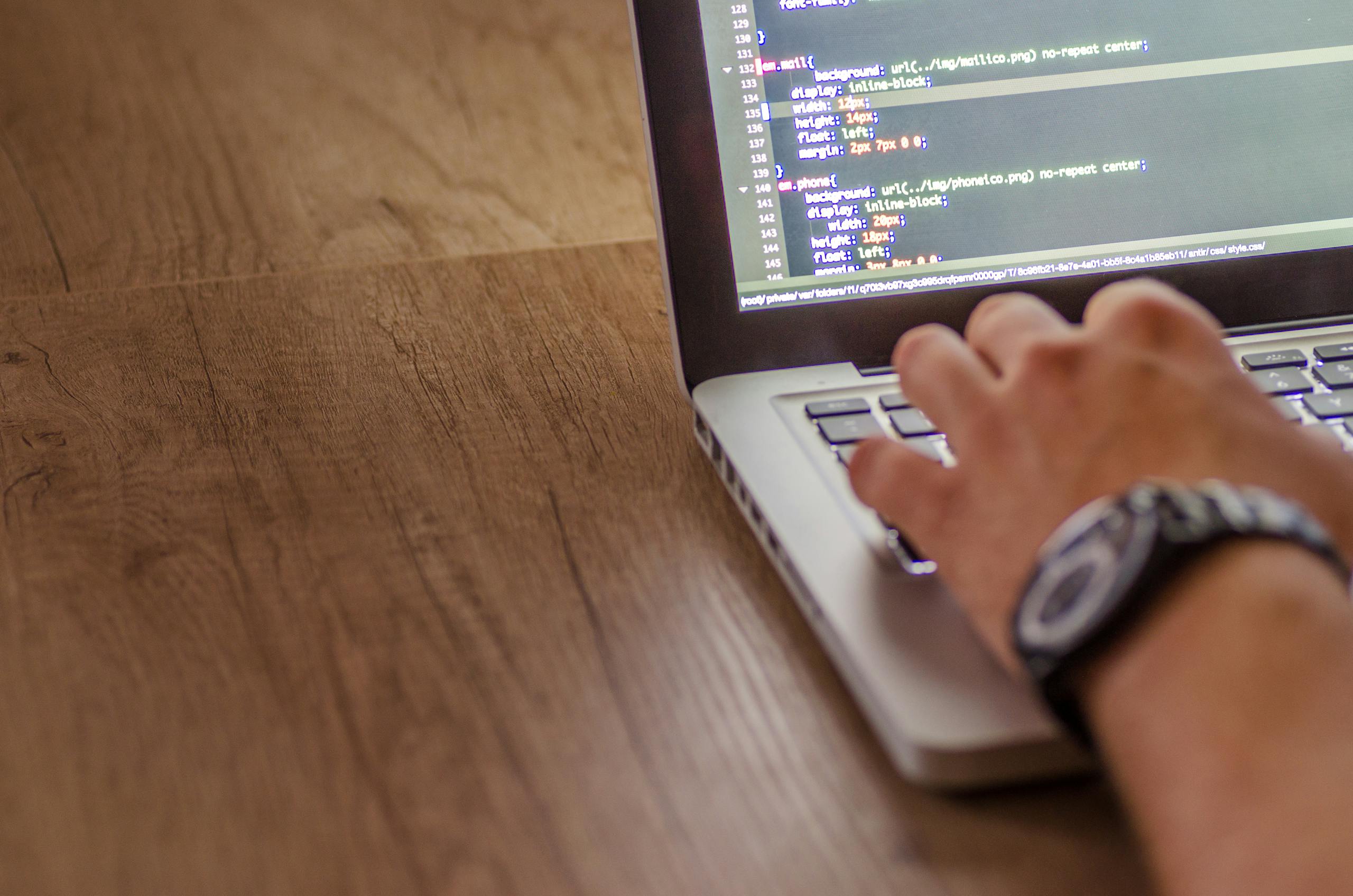 A person types on a laptop with code displayed on the screen at a wooden desk.